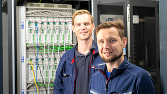 Zwei Männer in blauen Arbeitsjacken stehen vor einem geöffneten Serverrack mit grünen Kabeln